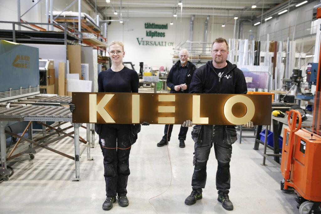 Kaksi henkilöä pitämässä isoa opastetta jossa lukee "kielo"