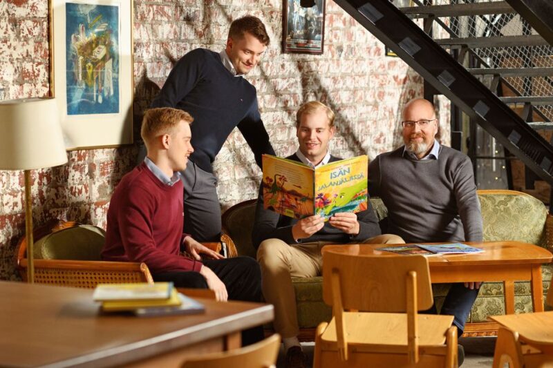 Atso Mäkelä istuu kolmen poikansa, Joonatanin, Samulin ja Santerin, kanssa kotoisasti sohvaryhmässä ja lukee kuvakirjaa, jonka nimi on "Isän jalanjäljissä".